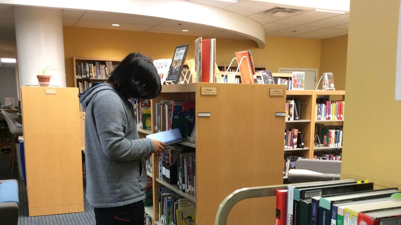A High School student, in profile, selects a book from a library bookshelf