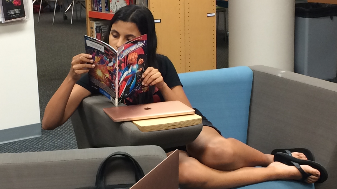 A High School girl in flip flops sits reading a comic book of a Latine superhero with her legs beside her